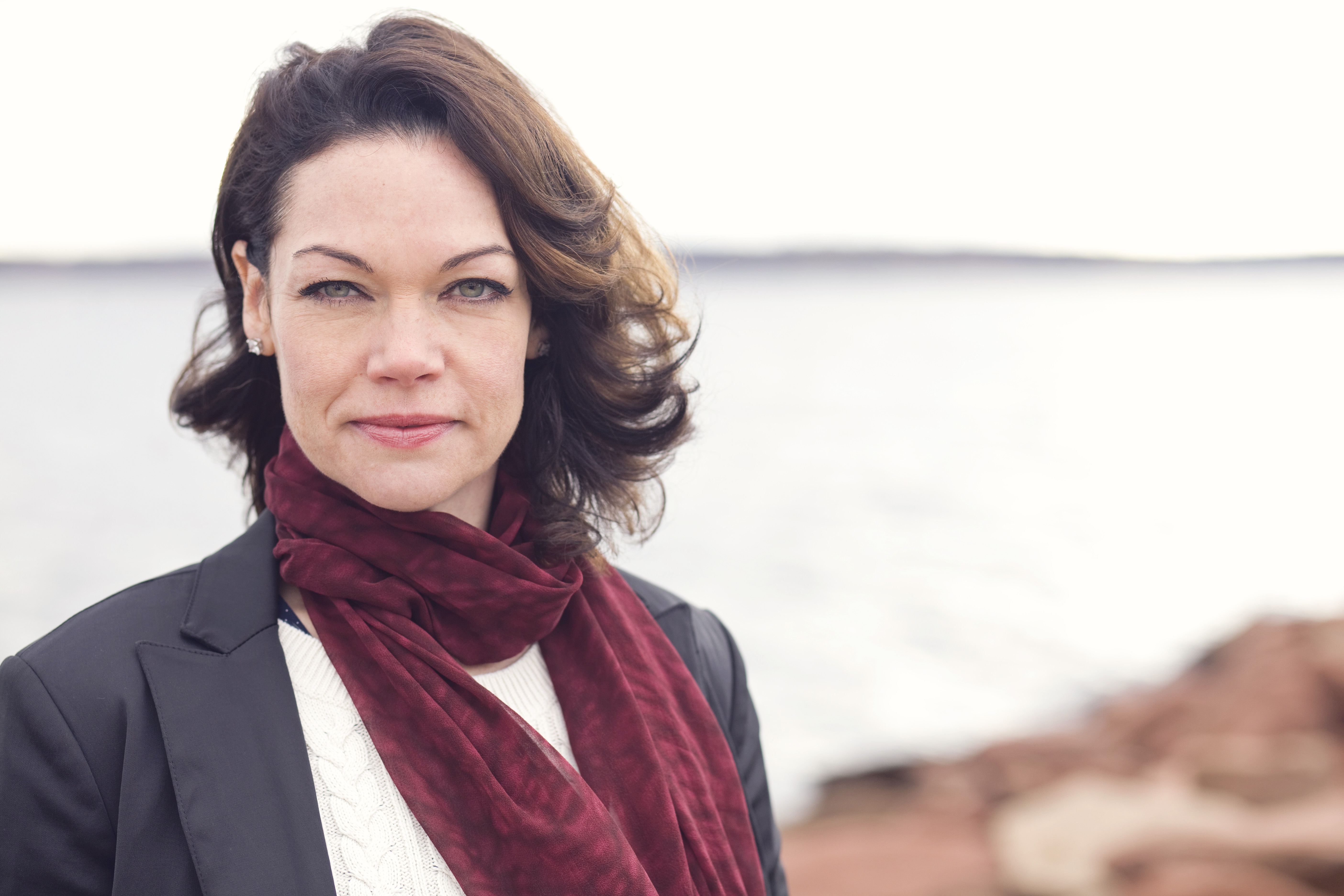 Jen standing on the beach wearing a white shirt and red scarf looking forward.